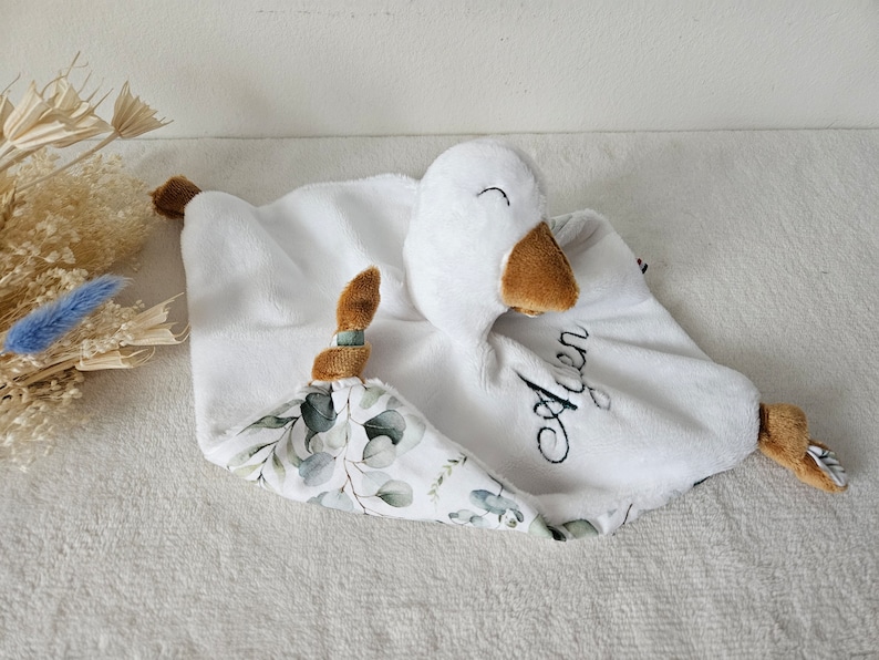May include: A white plush baby comforter with a goose head and brown beak and feet. The comforter has a white and green floral patterned corner and the name "Agen" embroidered in black.