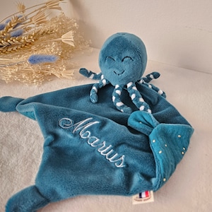 Peut inclure: Une couverture de sécurité en peluche bleu sarcelle avec une tête de pieuvre douce aux yeux et au sourire brodés. La couverture porte le prénom Marius brodé en écriture blanche, et un coin est orné de petits points dorés et d'une étiquette drapeau français.