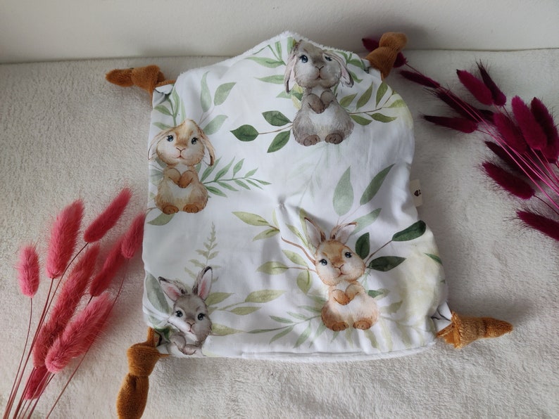 May include: A white baby security blanket with a pattern of watercolor bunnies and green leaves. The blanket has brown plush corners. The overall design is soft and inviting, perfect for a nursery or baby gift.