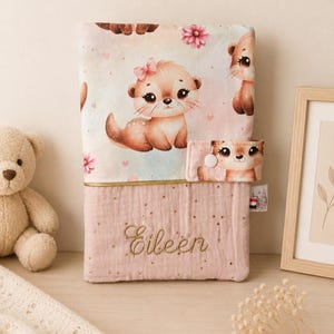 Pu&ograve; includere: Una copertina in tessuto rettangolare con una sezione superiore decorata da un motivo di lontre animate e fiori rosa. La parte inferiore &egrave; rosa polvere con puntini dorati e il nome Eileen ricamato. Presenta una chiusura a bottone automatico.