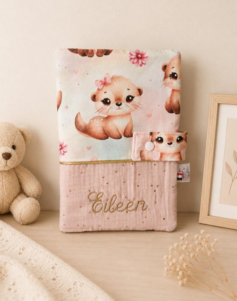 Pu&ograve; includere: Una copertina in tessuto rettangolare con una sezione superiore decorata da un motivo di lontre animate e fiori rosa. La parte inferiore &egrave; rosa polvere con puntini dorati e il nome Eileen ricamato. Presenta una chiusura a bottone automatico.