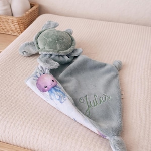 Peut inclure: Une couverture de sécurité en peluche vert sauge doux avec une tête et des nageoires de tortue en peluche sur le dessus. Le tissu est texturé et le prénom Jules est brodé en écriture cursive verte. Un coin présente une méduse violette imprimée.