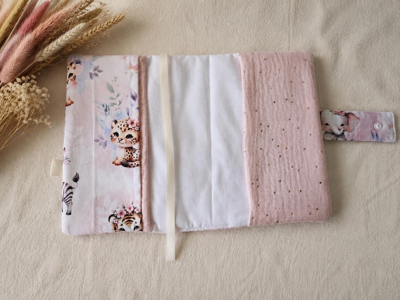 Pu&ograve; includere: Un fasciatoio rettangolare in tessuto con sfondo rosa chiaro e illustrazioni di cuccioli di animali tra cui un leopardo, una tigre e una zebra. Include un pannello centrale bianco, un pannello laterale rosa testurizzato con puntini dorati e una chiusura a pressione.
