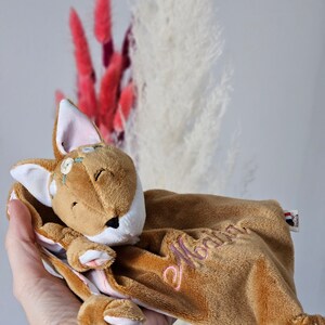 Peut inclure: Une doudou pour bébé en forme de renard en peluche marron avec des détails brodés. Le renard a les yeux fermés, des oreilles blanches avec l'intérieur rose et une couronne florale. Le doudou porte le nom "Marina" brodé en rose. Une petite étiquette est attachée.