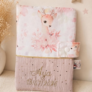 Pu&ograve; includere: Una copertina in tessuto rettangolare con una sezione superiore stampata con cerbiatti e fiori rosa e una sezione inferiore in rosa polvere con testo ricamato in oro Aja e 13/07/2026. Include una linguetta laterale con bottone a pressione.