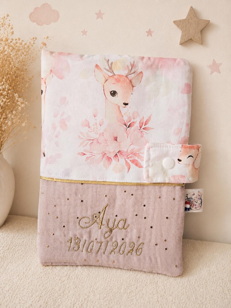Pu&ograve; includere: Una copertina in tessuto rettangolare con una sezione superiore stampata con cerbiatti e fiori rosa e una sezione inferiore in rosa polvere con testo ricamato in oro Aja e 13/07/2026. Include una linguetta laterale con bottone a pressione.