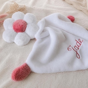 Peut inclure: Une couverture de sécurité en peluche blanche douce avec un accent en forme de fleur au centre rose et aux pétales blancs. La couverture présente des détails roses aux coins et le prénom Jade brodé en écriture cursive rouge sur le tissu.