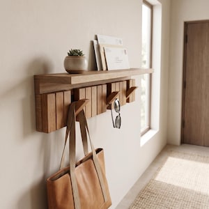 May include: A light brown wooden wall-mounted shelf featuring a series of vertical slats and integrated hooks. A small potted succulent sits on the top surface alongside several envelopes. A tan leather tote bag hangs from one hook.