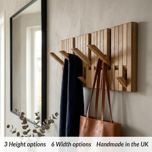May include: A wall-mounted wooden coat rack featuring vertical slats and multiple angled hooks. A dark blue scarf and a brown leather tote bag are hanging from the hooks. The text 3 Height options 6 Width options Handmade in the UK is visible.