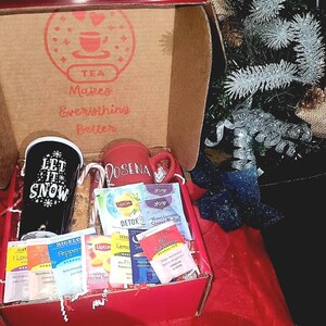 May include: A red and white gift box filled with tea bags and two mugs. The mugs are white with black lettering. One mug says "Let it snow" and the other says "Rosena". The box has a red lid with the words "Tea Makes Everything Better" printed on it.