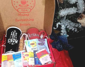 May include: A red and white gift box filled with tea bags and two mugs. The mugs are white with black lettering. One mug says "Let it snow" and the other says "Rosena". The box has a red lid with the words "Tea Makes Everything Better" printed on it.