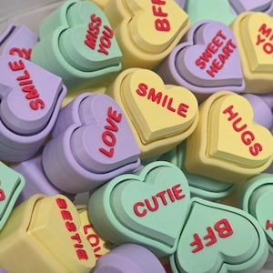May include: A collection of pastel-colored heart-shaped candies with various messages in red. The candies are in shades of yellow, green, and purple, with phrases like "Smile," "Love," and "BFF" displayed on them. These are likely Valentine's Day or friendship-themed treats.