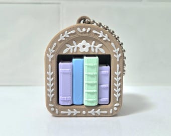 May include: A miniature bookshelf keychain with four colorful books. The frame is tan with white floral and leaf accents. The books are pastel purple, blue, and green. A silver chain is attached for hanging.
