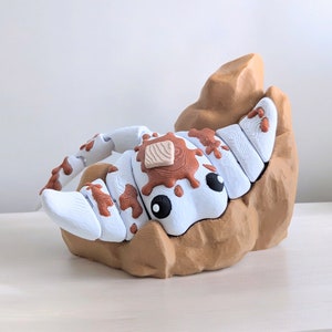 May include: A white and brown 3D printed toy shaped like a scorpion, with a brown patch on its back, sitting on a brown rock.