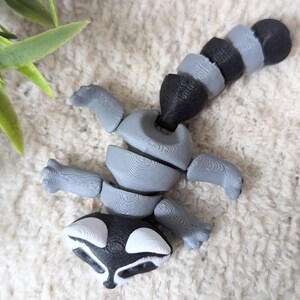 Peut inclure: Une figurine articul&eacute;e de raton laveur gris et noir avec une queue segment&eacute;e. La figurine est en plastique et a un visage d&eacute;taill&eacute;.