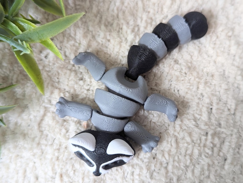 Peut inclure: Une figurine articul&eacute;e de raton laveur gris et noir avec une queue segment&eacute;e. La figurine est en plastique et a un visage d&eacute;taill&eacute;.
