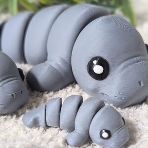 May include: Three gray 3D printed manatee figurines. The largest manatee is lying down with its head turned to the side. The smallest manatee is curled up. The middle-sized manatee is lying on its side with its head turned to the side.
