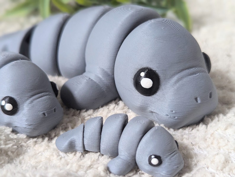 May include: Three gray 3D printed manatee figurines. The largest manatee is lying down with its head turned to the side. The smallest manatee is curled up. The middle-sized manatee is lying on its side with its head turned to the side.