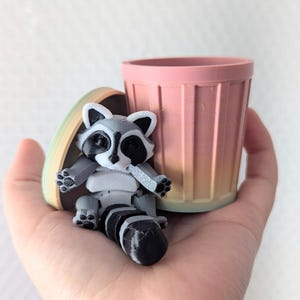 Peut inclure: Une petite figurine de raton laveur gris et blanc assise dans une poubelle rose et orange d&eacute;grad&eacute;e. La poubelle est en plastique et a une texture nervur&eacute;e.