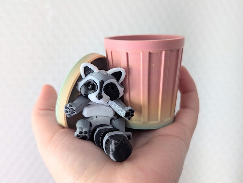Peut inclure: Une petite figurine de raton laveur gris et blanc assise dans une poubelle rose et orange d&eacute;grad&eacute;e. La poubelle est en plastique et a une texture nervur&eacute;e.