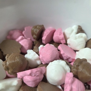 May include: Close-up of a collection of decorative, stylized acorn-shaped objects. The acorns are in shades of pink, white, and brown. The objects have a textured, detailed surface, and are clustered together in a white container.