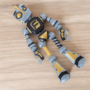 May include: A silver and gold robot toy with black accents. The robot has a large head with a gold faceplate and a black letter "F" on its chest.