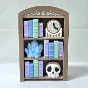 May include: A miniature brown bookshelf with three shelves. The shelves hold colourful books, a clock, a blue crystal, and a white skull. The books are in shades of purple, blue, and green. The clock has a white face and black hands.