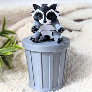 Peut inclure: Une petite poubelle grise avec une figurine de raton laveur assise dessus. Le raton laveur est noir et blanc avec un ventre blanc et est assis avec ses pattes tendues.