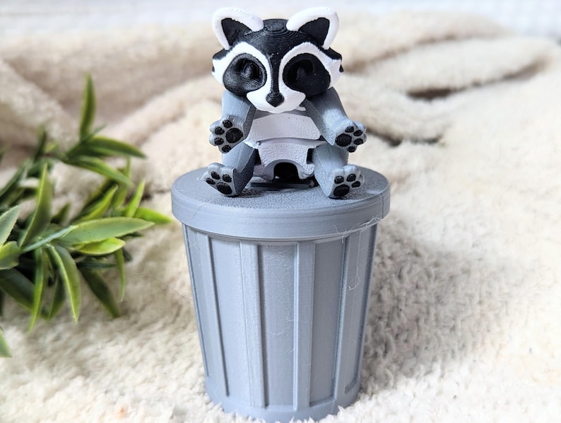 Peut inclure: Une petite poubelle grise avec une figurine de raton laveur assise dessus. Le raton laveur est noir et blanc avec un ventre blanc et est assis avec ses pattes tendues.