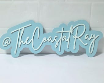 May include: A light blue sign with the text "@ The Coastal Ray" in white script. The sign is in a cloud-like shape and is likely a decorative item for a coastal-themed space.