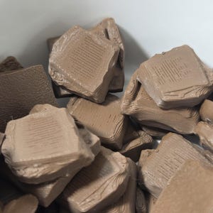 May include: Close-up of a pile of light brown, irregularly shaped objects. The objects appear to be stacked and have a textured surface with visible lines. The overall impression is of a collection of small, blocky forms.