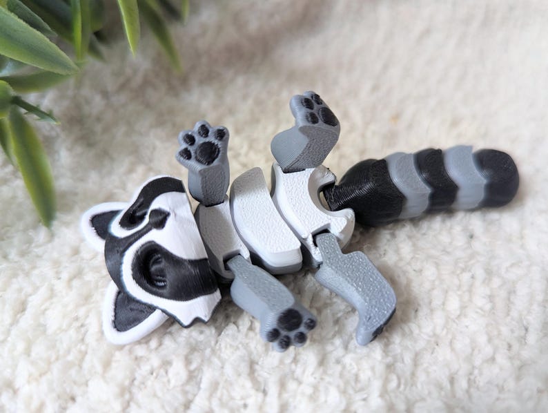 Peut inclure: Une figurine articul&eacute;e de raton laveur en plastique blanc et noir avec un corps segment&eacute; et une queue enroul&eacute;e. La figurine a un design r&eacute;aliste.