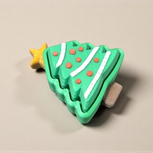 May include: A green Christmas tree-shaped hair clip with white accents and orange dots. A yellow star is on the left side. The clip is made of plastic and is a festive accessory.