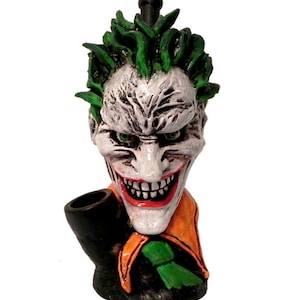 May include: A ceramic Joker head pipe with green hair, white skin, red lips, and a wide grin. The pipe has a black bowl and an orange and green collar.