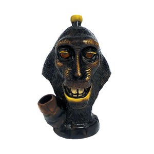 May include: A dark brown monkey-faced sculpture with a pipe. The sculpture has a smiling expression with visible teeth and orange eyes. A small yellow cylinder is on top of the head. The pipe is brown.