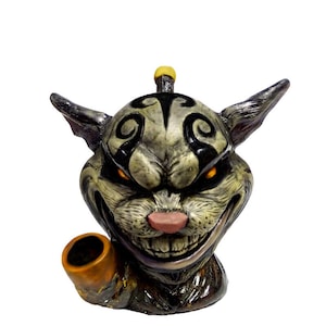 May include: A ceramic pipe shaped like a snarling, grey and black cat with glowing red eyes. The pipe has a brown, cylindrical bowl.