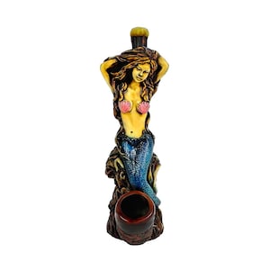 May include: A mermaid-shaped pipe with a brown, yellow, and blue painted design. The mermaid is wearing a pink seashell bra and a blue tail. The pipe has a brown bowl.