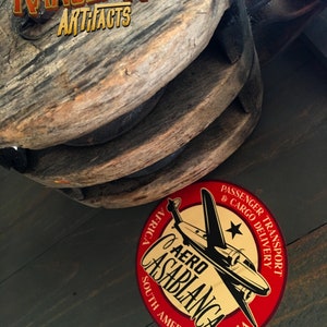 May include: A red and gold round sticker with the text "AERO CASABLANCA" and a black and white illustration of an airplane. The sticker also includes the text "PASSENGER TRANSPORT & CARGO DELIVERY" and the words "AFRICA, SOUTH AMERICA, INDIA".