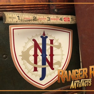 May include: A vintage-style sticker with a compass rose and the letters "NJ" in red, blue, and gold. The sticker is on a dark green surface and has the text "RANGER ROB ARTIFACTS" below it.