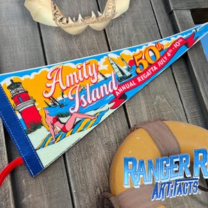May include: A triangular pennant with the words "Amity Island" and "50th Annual Regatta July 4th-10th" in a vintage style. The pennant features a lighthouse, sailboats, and a woman sunbathing. A life preserver with "Ranger Rob Artifacts" is also visible.