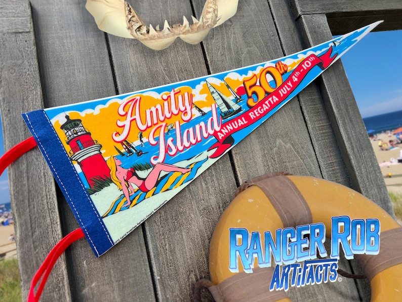 May include: A triangular pennant with the words "Amity Island" and "50th Annual Regatta July 4th-10th" in a vintage style. The pennant features a lighthouse, sailboats, and a woman sunbathing. A life preserver with "Ranger Rob Artifacts" is also visible.