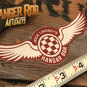 May include: A red and white sticker with a checkered circle and wings. The text on the sticker reads "Jock Lindsey's Hangar Bar".