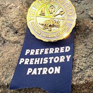 May include: A gold-colored medallion from The Dino Institute with a triceratops design, attached to a blue ribbon that reads "Preferred Prehistory Patron". The medallion has the words "Exploration Excavation Exubation" around the edge.