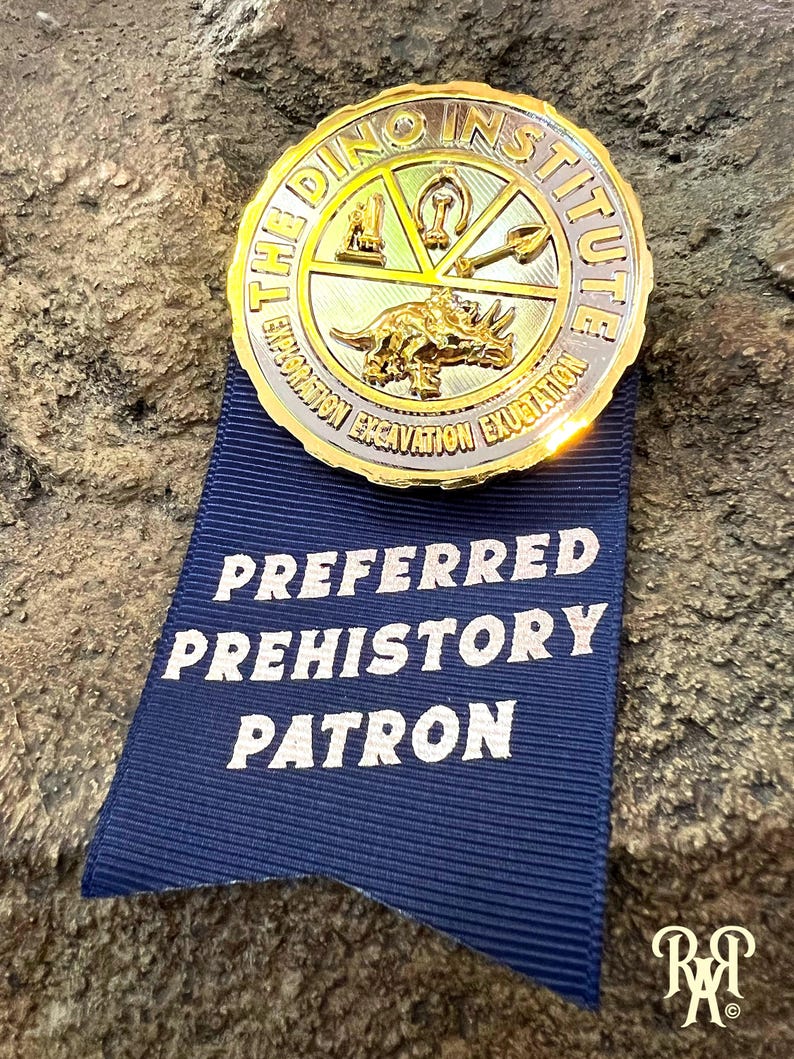May include: A gold-colored medallion from The Dino Institute with a triceratops design, attached to a blue ribbon that reads "Preferred Prehistory Patron". The medallion has the words "Exploration Excavation Exubation" around the edge.