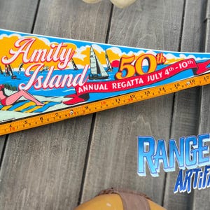May include: A pennant-shaped banner with the words "Amity Island" and "50th Annual Regatta July 4th-10th." The banner features a lighthouse, sailboats, and a woman sunbathing. A ruler is along the bottom.