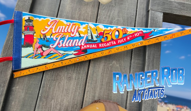 May include: A pennant-shaped banner with the words "Amity Island" and "50th Annual Regatta July 4th-10th." The banner features a lighthouse, sailboats, and a woman sunbathing. A ruler is along the bottom.