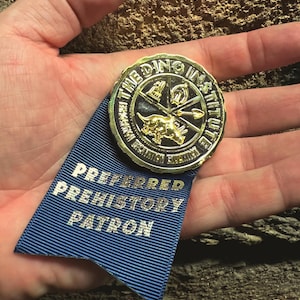 May include: A gold-colored medallion with the text "THE DINO INSTITUTE" and a dinosaur image, attached to a blue ribbon. The ribbon reads "PREFERRED PREHISTORY PATRON" in silver lettering. The image is set against a textured background.