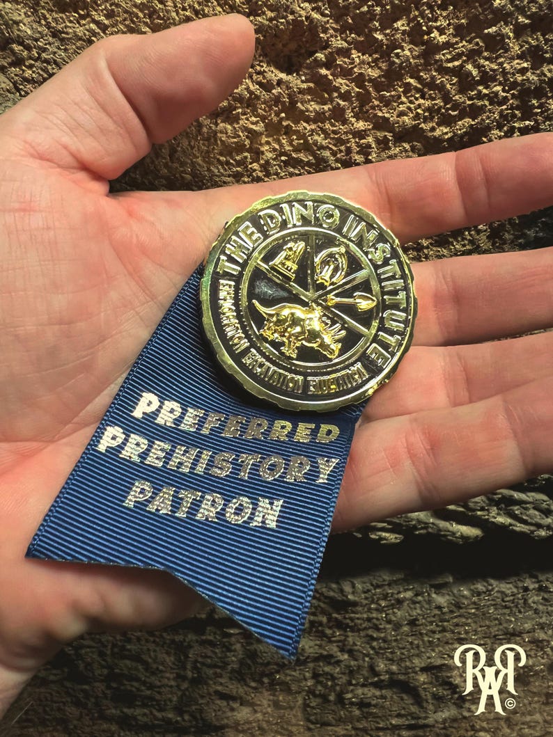 May include: A gold-colored medallion with the text "THE DINO INSTITUTE" and a dinosaur image, attached to a blue ribbon. The ribbon reads "PREFERRED PREHISTORY PATRON" in silver lettering. The image is set against a textured background.