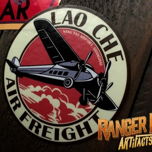 May include: A vintage-style round sticker with a red, white, and black design. It features a plane flying through the clouds with the text "Lao Che Air Freight" and "Nang Tao Airport, Shanghai".
