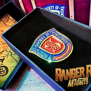 May include: A gold-colored pin with a blue and red shield design. The shield features a snake with a crown and the text "Society of Explorers & Adventurers - Explorations Continue". The pin is in a black velvet box with the text "Ranger Rob Artifacts".